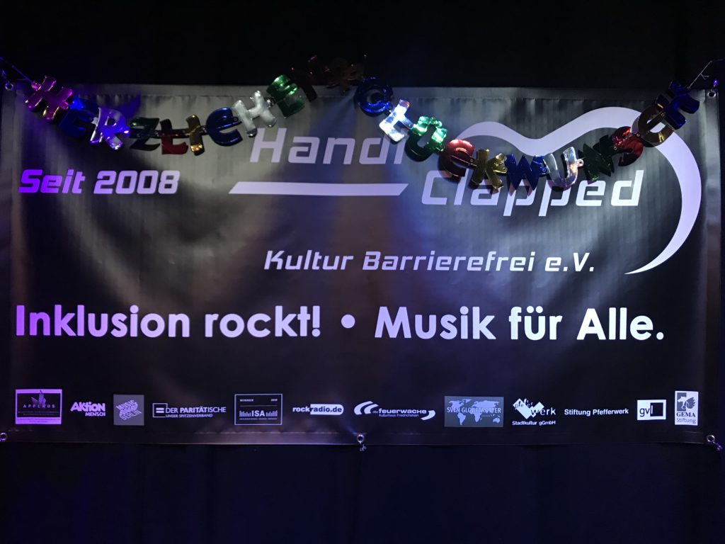 Banner auf einem Konzert, darauf steht Handiclapped - Kultur Barrierefrei e.V. Inklsuion rockt! Musik für Alle.